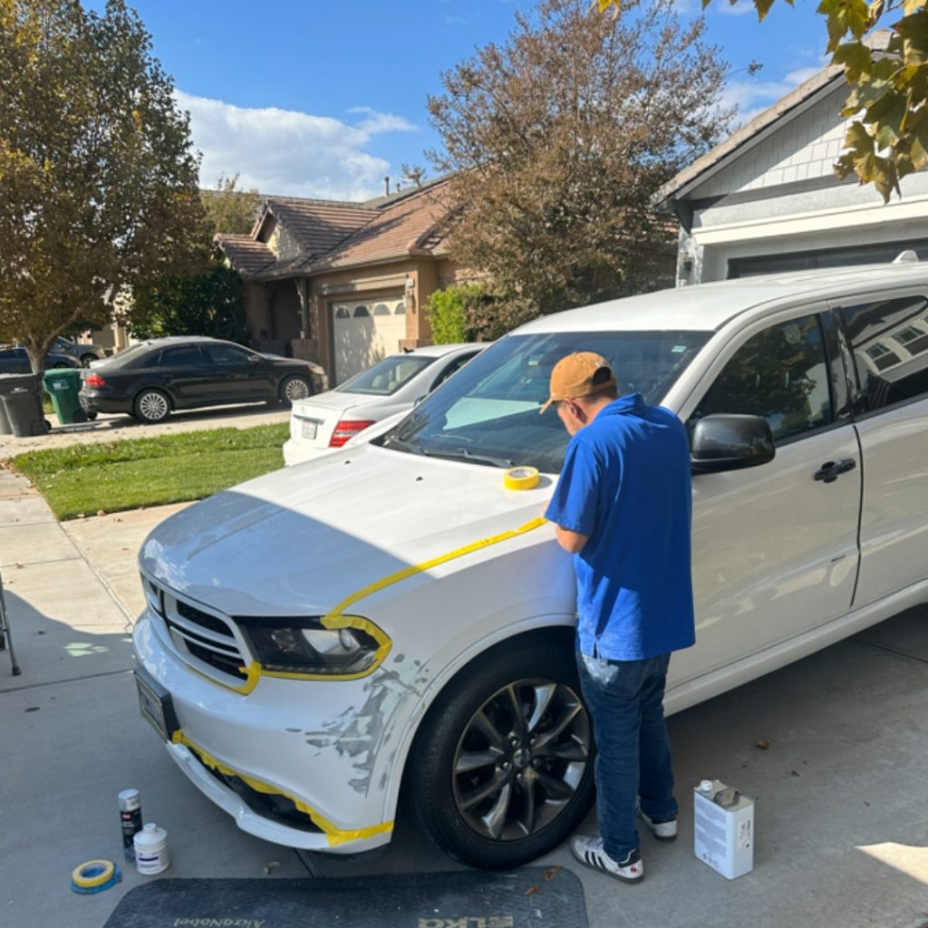 Mobile technician in blue shirt and yellow cap inspecting white sedan with yellow tape marking damage on hood and bumper in residential driveway